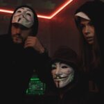 People wearing Guy Fawkes masks in a dark technology room, representing anonymous cyber criminals and concealed threat actor identities.