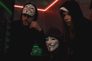 People wearing Guy Fawkes masks in a dark technology room, representing anonymous cyber criminals and concealed threat actor identities.