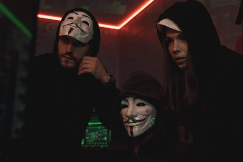 People wearing Guy Fawkes masks in a dark technology room, representing anonymous cyber criminals and concealed threat actor identities.