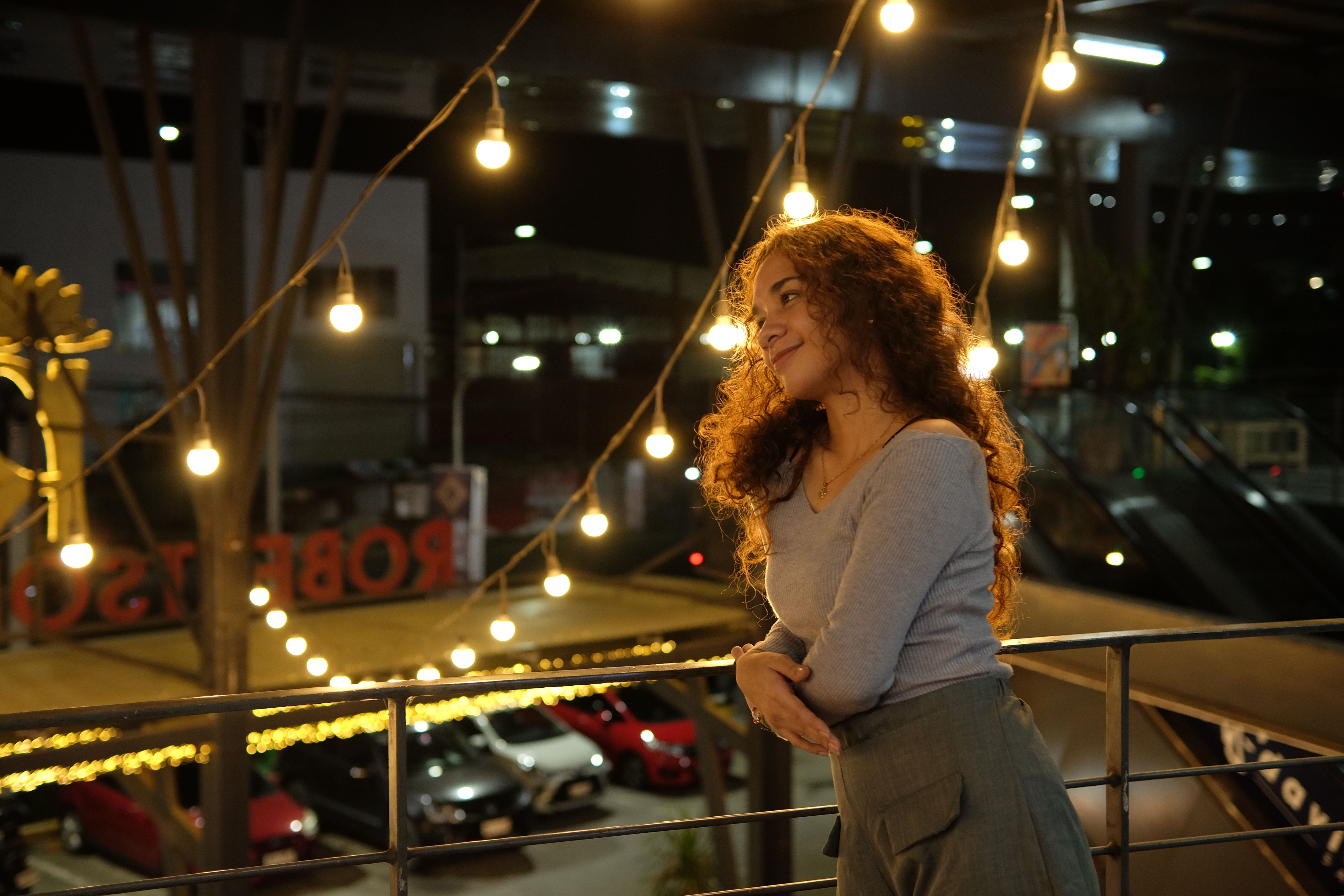 A woman on a balcony at night, lit by warm string lights